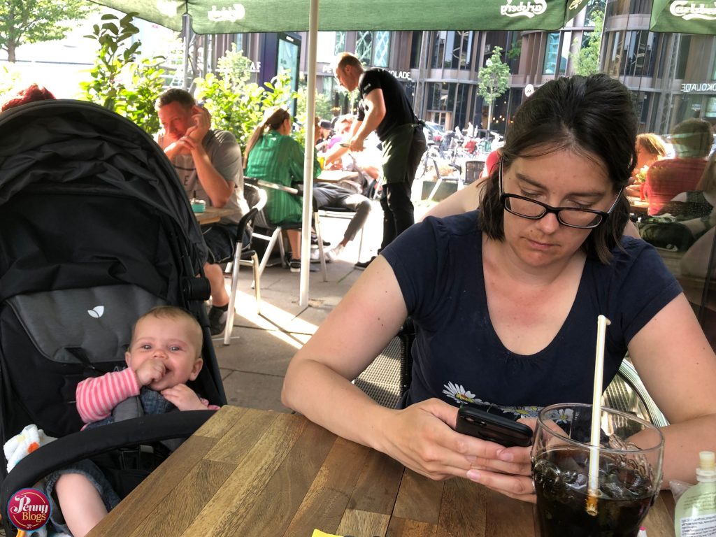 Penny sat using her phone in a restaurant in Copenhagen with Tube Stop Baby sat next to her in her pushchair smiling. Photo to accompany an article on the Three App.