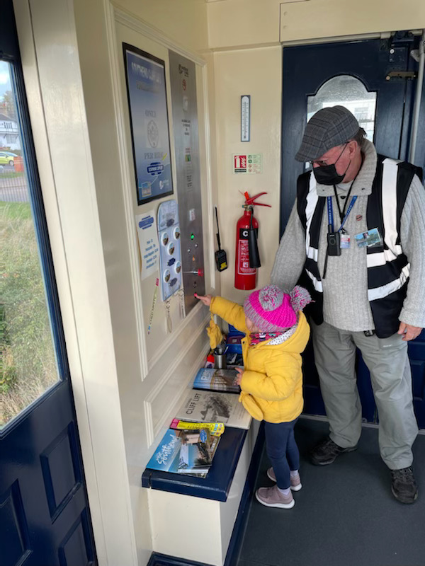 A two year old girl wearing a yellow coat and pink bobble hat is pressing the button to operate the lift whilst under the watchful eye of one of the volunteers who is stood next to her wearing a jacket with reflective stripes, a lanyard round his neck, a black face mask and a flat cap.