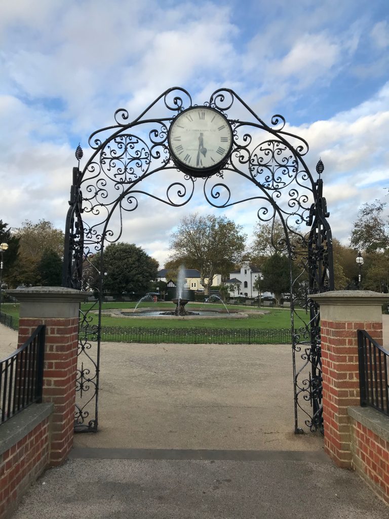 Looking through the orange gateway into Prittwell Gardens in Southend-on-sea. You are looking at an ornate fountain in the middle of the gardens, and there is a clock (showing the wrong time!) above the entrance as part of the ornate metal gateway.
