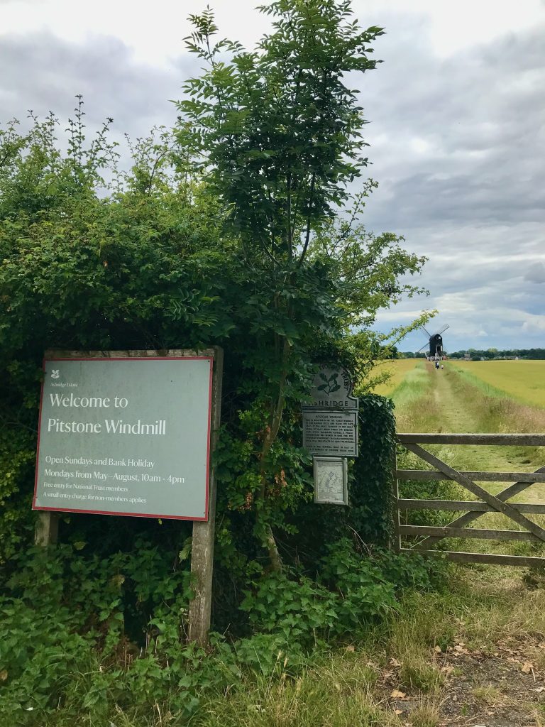 A view across fields looking towards Pitstone Windmill. The windmill is in the distance and there is a grass path leading to it. In the foreground is a National Trust sign welcoming visitors to the windmill.