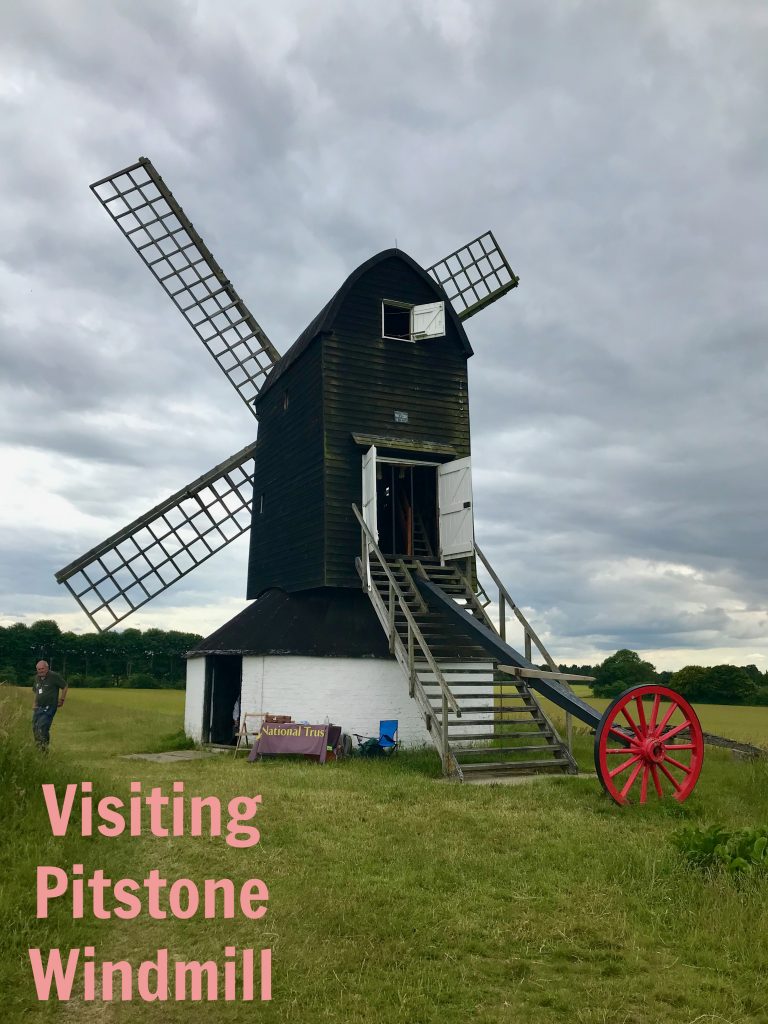 Looking at the rear of Pitstone Windmill. You can see teh ends of teh sails on teh front of teh windmill and the stairs leading up into the windmill and the wooden arm with the turning wheel on the end of it. Also in picture is teh National Trust welcome table outside the windmill. Text has been added to the photographs saying "Visiting Pitstone Windmill".