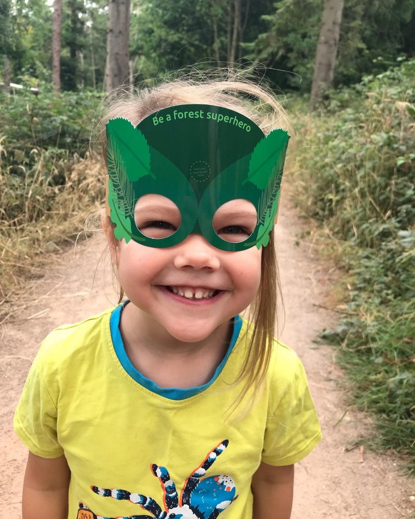 A close up of a smiling three year old girl with blonde hair and wearing a yellow t-shirt. On her face is a paper forest superhero mask that she got in her Superworm trail activity pack.
