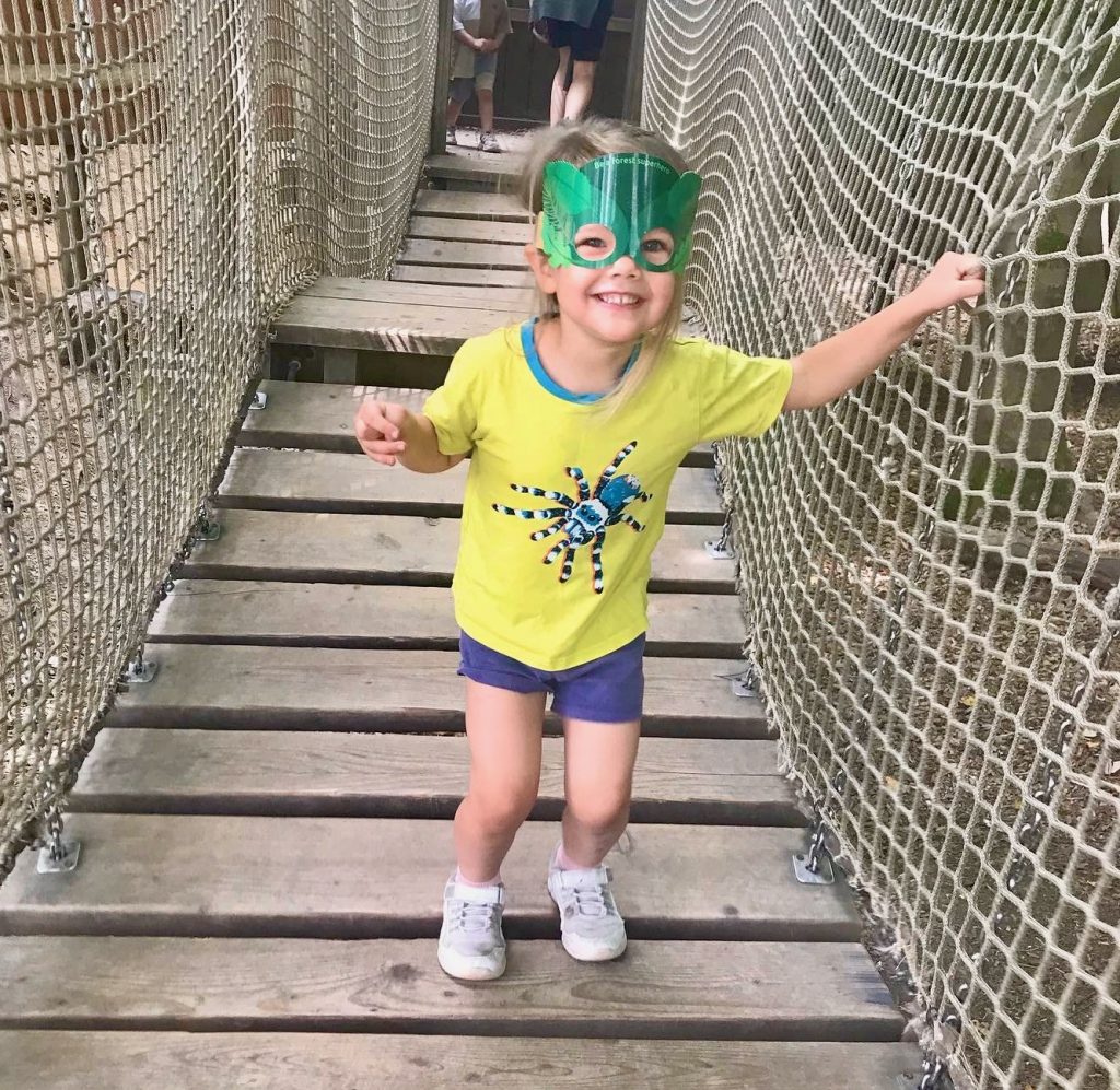 A smiling three year old girl wearing a yellow t-shirt and navy shorts. She also has on a paper "forest Superhero" face mask. She is stood on a wobbly bridge with netting on both sides of her.