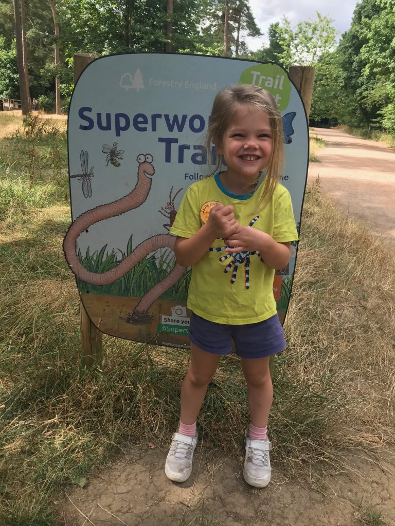 A smiling three year old with blonde hair stood in front of a sign for the Superworm trail. She is wearing a yellow t-shirt and navy shorts.