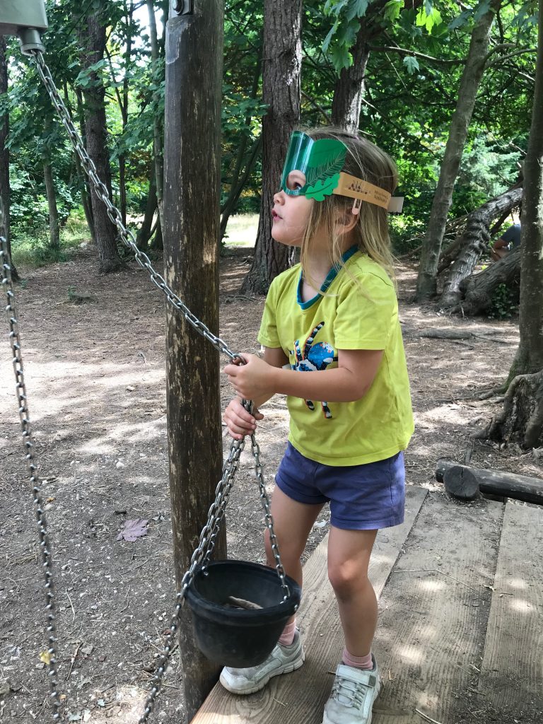 A three year old girl wearing a yellow t-shirt and navy shorts and a forest superhero face mask. She is in a wooden playground and is hold ing a metal chain that is attached to a rubber bucket. She looks like she is hauling the bucket up using the chain.