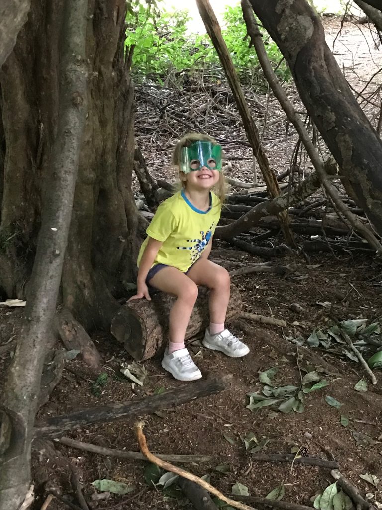 A smiling three year old girl wearing a yellow t-shirt, navy shorts and a forest superhero face mask sitting on a log in a wooden den.