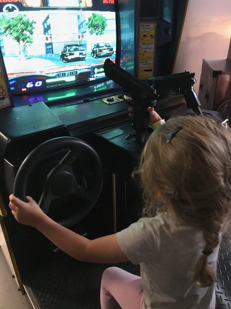 A picture of a three year old girl sat at an arcade machine. On the screen is a driving game and she has her left hand on a steering wheel and her right hand on a gun.