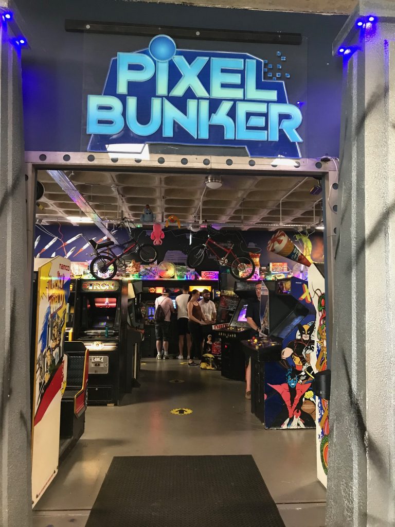 A view into the Pixel Bunker in Milton Keynes. You are looking into the doorway and above it is a large illuminated sign saying The Pixel Bunker. Inside you can see some unidentified people in a dark room filled with arcade games.