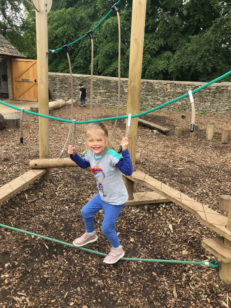 A smiling four year old girl wearing blue leggings and a long sleeved t-shirt  balancing on a tightrope whilst holding on to some rope supports.