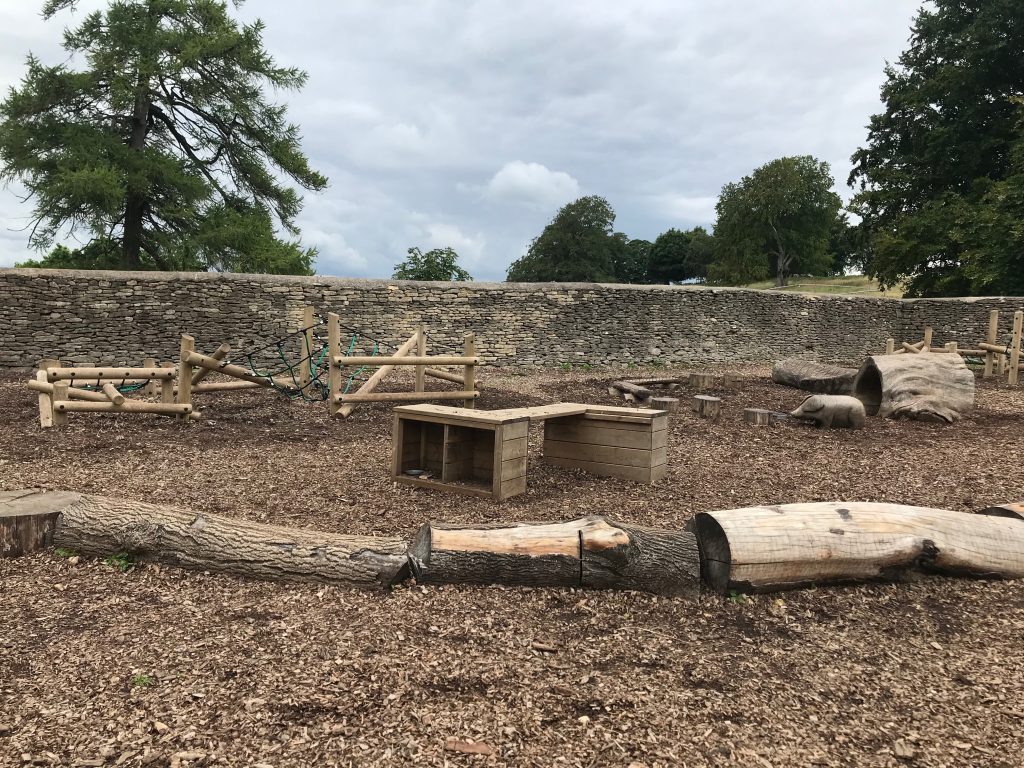A wooden playground featuring a variety of balancing and climbing opportunities for young children and a wooden pig. All surrounded by a bark chipping ground covering. 