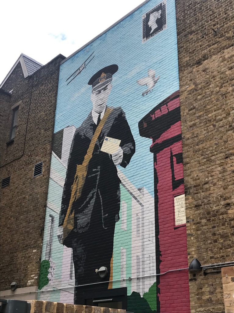 An image painted onto the side of a brick building showing an old fashioned Postman holding a letter which it looks like he is about to place into a post box. You can see a bi-plane flying in the sky in the picture and in the top right hand corner is a Penny Black Postage Stamp.
