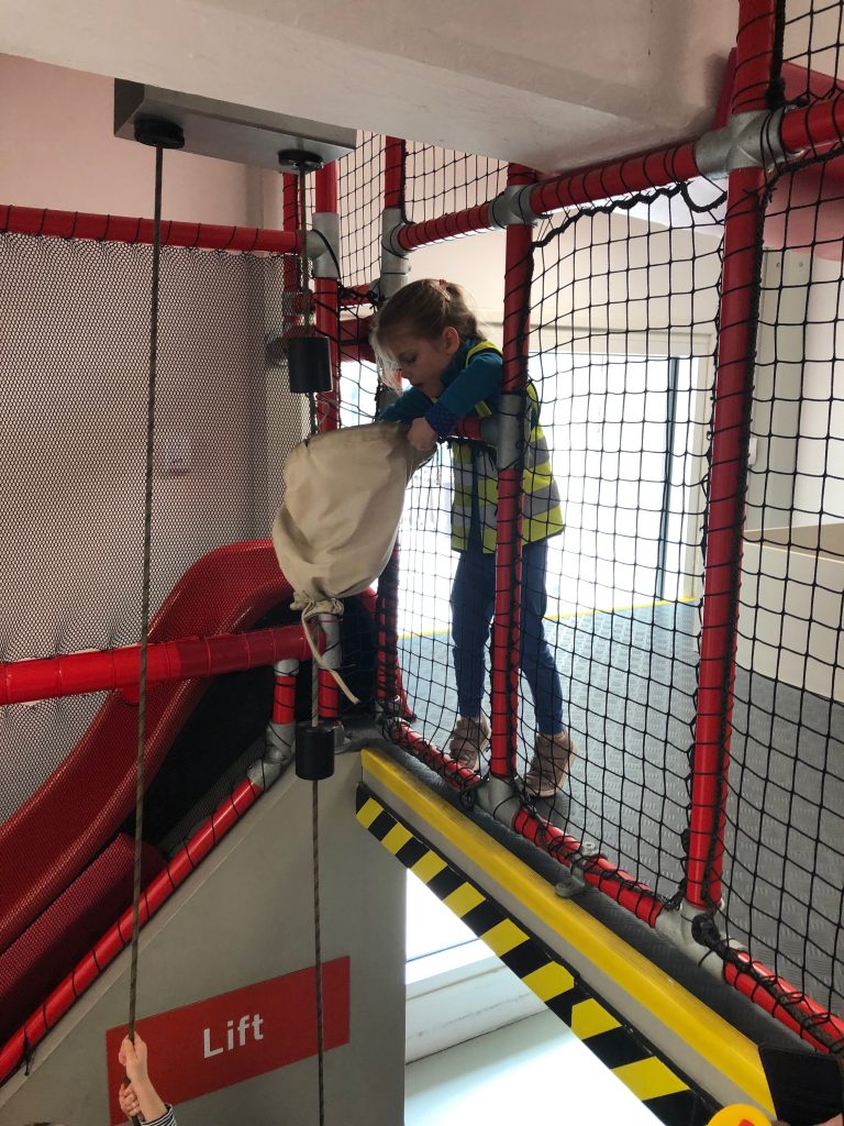A four year old girl wearing a child sized Royal Mail high-via jacket is hauling a play postal sack up after it has been raised to first floor level on a pulley system. Just next to where she is doing this you can see a red slide leading downstairs.