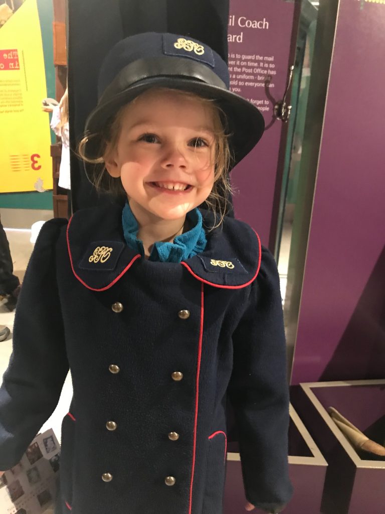 A blonde four year old girl smiling at the camera whilst wearing a vintage style GPO uniform of coat and hat, remade in a child's size.