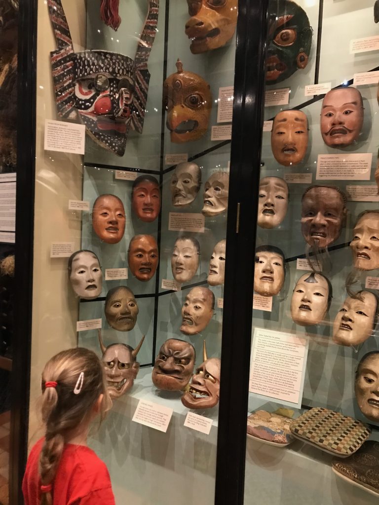 A display case full of slightly creepy looking masks with a four year old girl looking on.