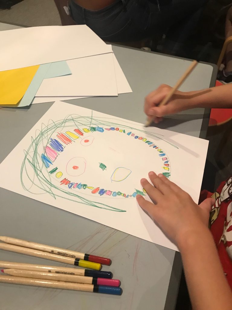 The hands of a four year old child as she draws a brightly coloured mask with coloured pencils.