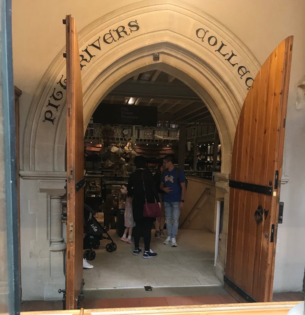A double wooden door set into a stone wall. Through teh door you can just see some museum display cases. Above the door are the words "Pitt Rivers Collection" and this door leads from teh maim hall of teh Oxford University Museum of Natural History to the Pitt Rivers Museum.