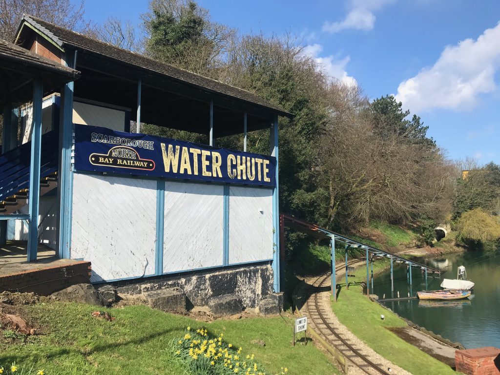 A view showing the timber building from which the water chute is launched and the metal track leading down into the lake. You can see a railway track passing under the metal track of the water chute. On the side of the building is a large banner reading "Scarborough North Bay Railway Water Chute"