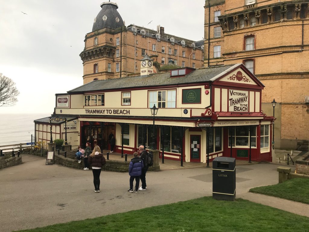 A picture showing the top station. The long building has a small clocktower on top, is mainly wooden in construction painted in cream and Burgundy. The words Tramway to Beach are along the side of the building and at the end it says Victorian Tramway to the beach.