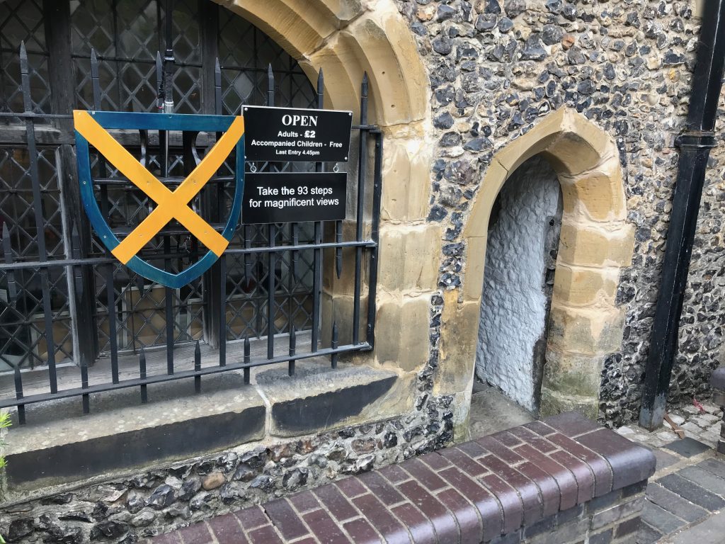 A small open doorway into a flint building. On teh railings are a large copy of the St Albans shield and a sign saying that you can climb the 93 steps to the top and what it costs.