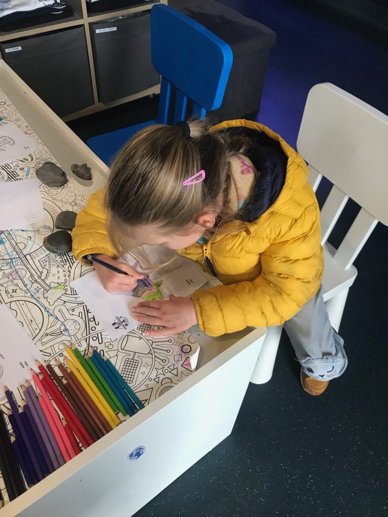 A five year old girl in a yellow coat sat at a small white table colouring in a picture of a fish.
