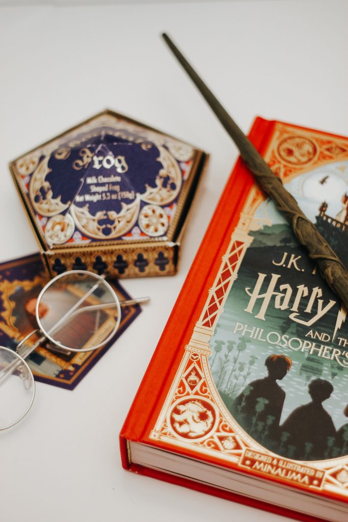 A top down photograph showing a copy of Harry Potter and the Philosopher's Stone, a pair of Harry's iconic glasses and a Chocolate Frog box.