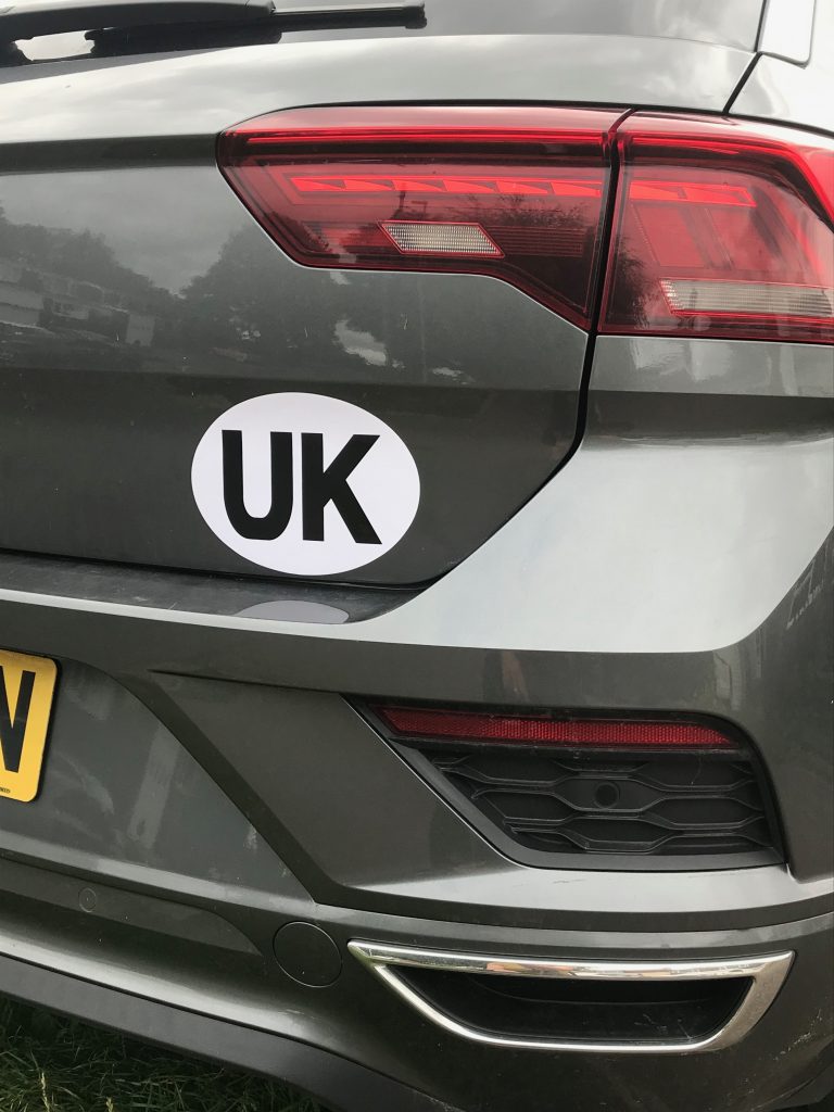 A image showing pat of the back of a car featuring a light cluster and an oval white sticker with "UK" on it in black. You can just catch a glimpse of the last letter on a yellow number plate on the edge of the photo.
