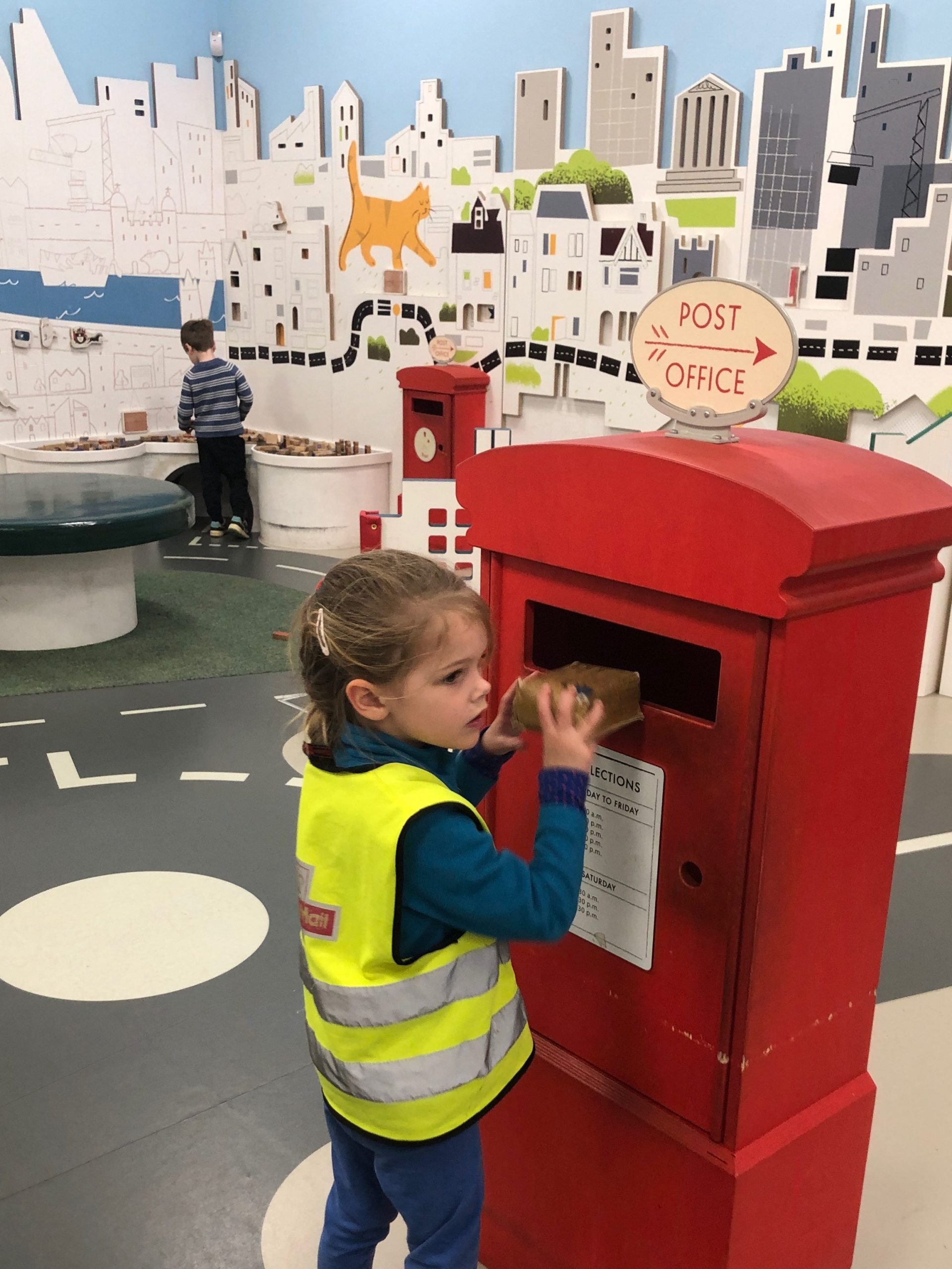 A 4 year old girl posts a parcel into a play post box at the Postal Museum's Sorted Role play area. The girl is wearing a Royal Mail high coz jacket in a child's size.